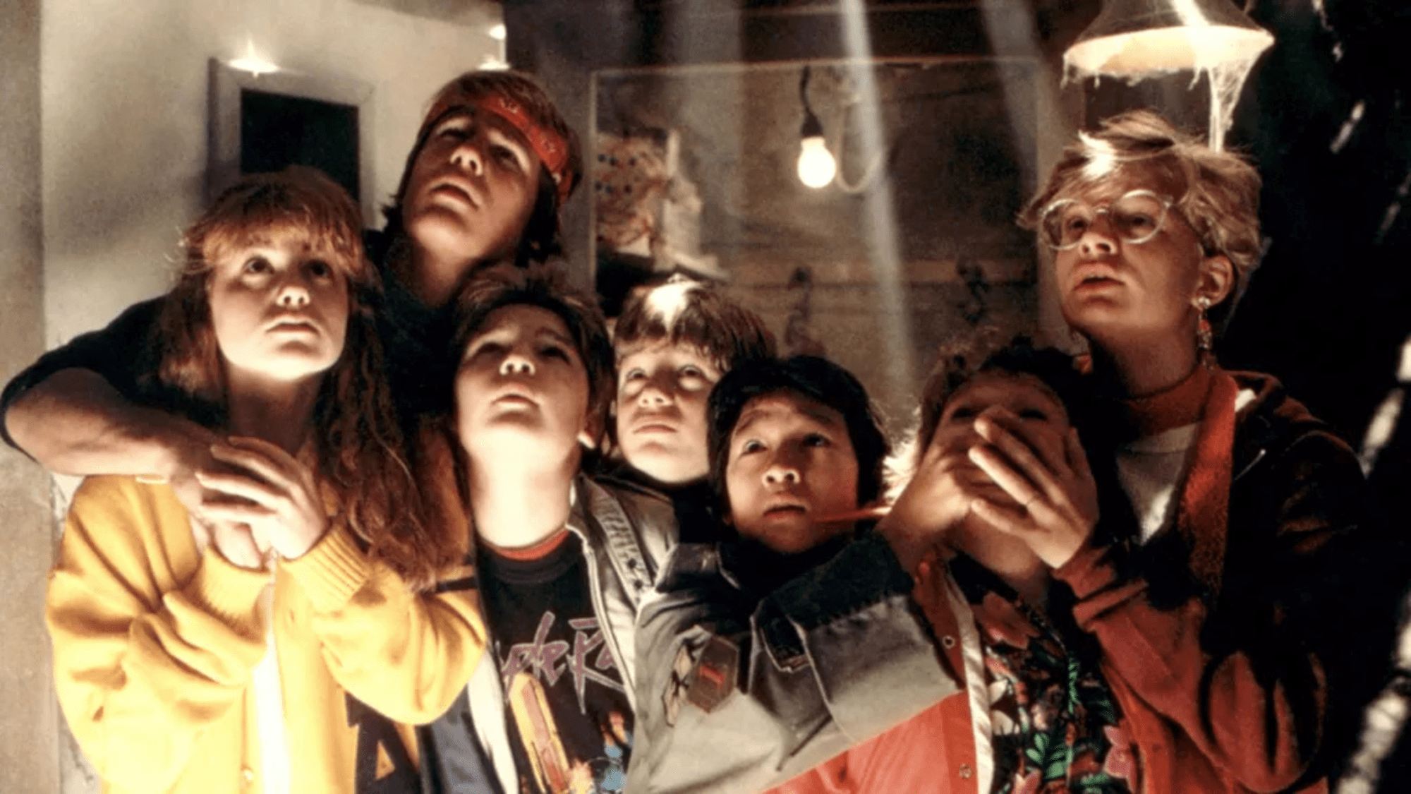 A group of children huddles closely together, gazing upward with expressions of awe and apprehension, illuminated by a single hanging lightbulb in a dim, cobweb-filled room.